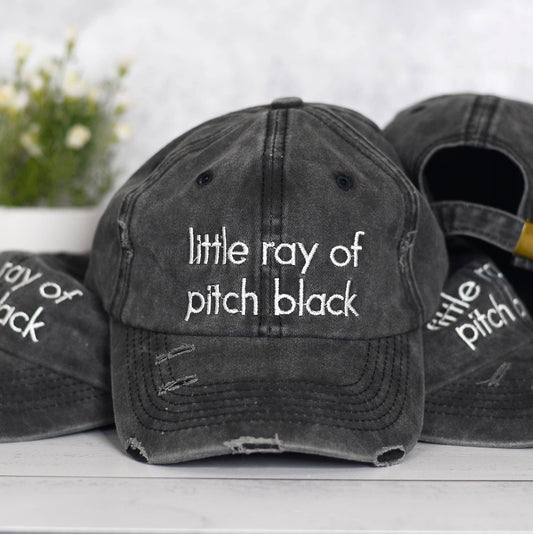 Black distressed baseball cap with 'little ray of pitch black' text on a white surface.